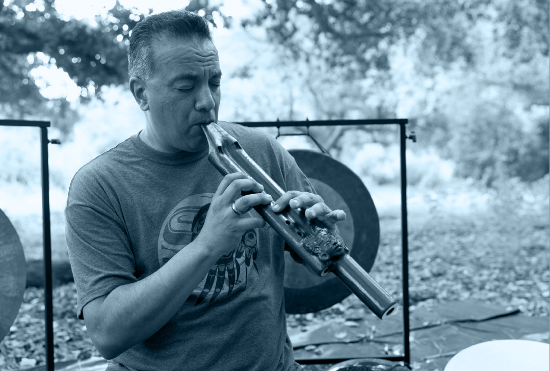 Gene Jimenez playing a wooden flute with gongs behind him